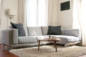 Sectional couch with light grey upholstery, freshly cleaned, windows in background and coffee table in front. White rug underneath couch