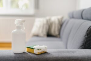 White spray bottle on the arm of a couch, a sponge with suds is next to the bottle