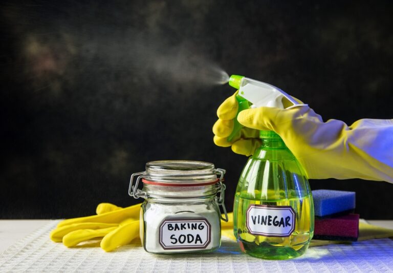 Gloved hand spraying a bottle with a vinegar label. A container of baking soda is to the left, sponges in the back